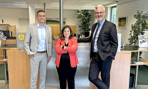 Drei Personen vor dem Bankschalter der Volksbank-Geschäftsstelle Brüchermühle: Tom Schwanbeck (links), Anna-Lena Zauner (Mitte), Sebastian Bender (rechts).