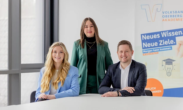 Isabell Stahl steht hinter Laura Harscheid und Mirko Müller, die nebeneinander sitzen. Alle drei blicken freundlich in die Kamera.