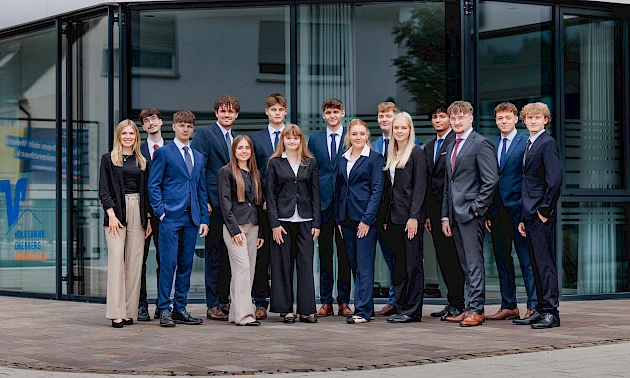 Gruppenfoto vor der Hauptstelle der Volksbank Oberberg in Wiehl. Die 14 neuen Auszubildenden stehen zusammen mit Ausbildungsleiterin Sarina Scherbaum (links) und blicken freundlich in die Kamera.