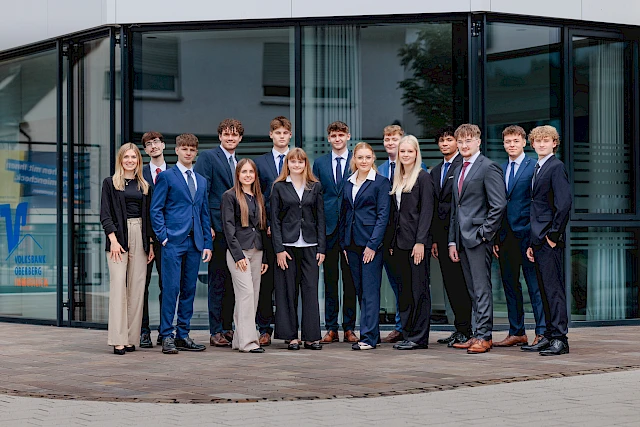 Gruppenfoto vor der Hauptstelle der Volksbank Oberberg in Wiehl. Die 14 neuen Auszubildenden stehen zusammen mit Ausbildungsleiterin Sarina Scherbaum (links) und blicken freundlich in die Kamera.