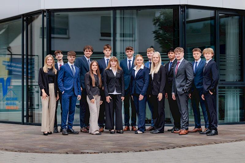 Gruppenfoto vor der Hauptstelle der Volksbank Oberberg in Wiehl. Die 14 neuen Auszubildenden stehen zusammen mit Ausbildungsleiterin Sarina Scherbaum (links) und blicken freundlich in die Kamera.