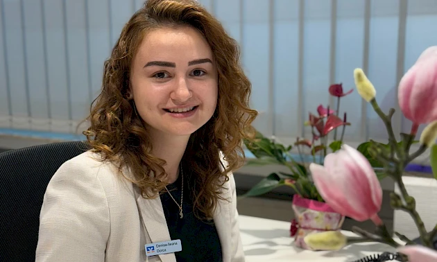 Auszubildende Denisa Dorca sitzt an ihrem Arbeitsplatz in der Geschäftsstelle Bielstein und lächelt in die Kamera.