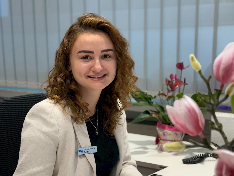 Auszubildende Denisa Dorca sitzt an ihrem Arbeitsplatz in der Geschäftsstelle Bielstein und lächelt in die Kamera.