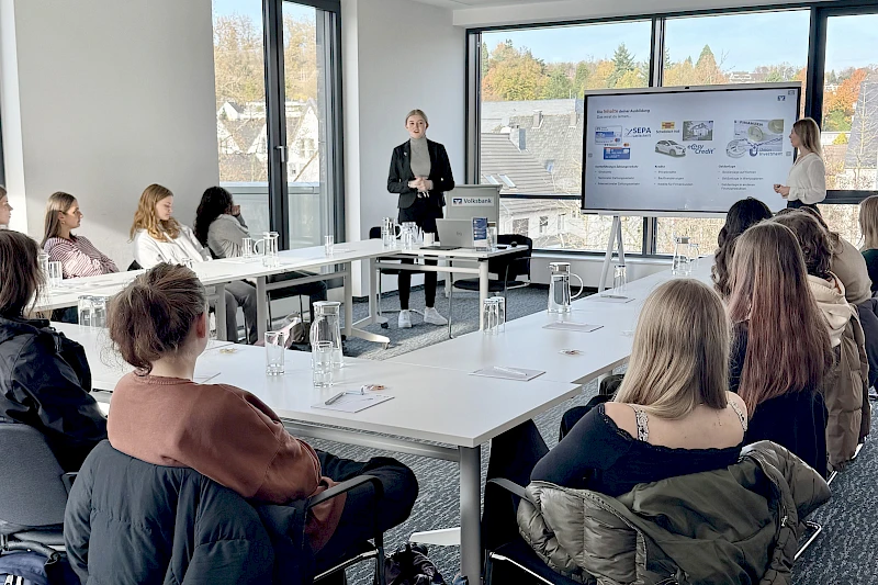 Vortragssituation in der Volksbank Oberberg Akademie: In einem großen Konferenzraum sitzen Schülerinnen und Schüler in U-Form an Tischen, während Sarina Scherbaum und Annabell Strauch vorne stehen und den Jugendlichen das Ausbildungsangebot der Volksbank