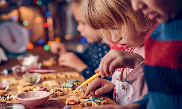 Mehrere Kinder sitzen an einem Tisch und verzieren gemeinsam Weihnachtsplätzchen mit Zuckerguss und Streuseln, im Hintergrund leuchten bunte Lichter und sorgen für eine warme, weihnachtliche Atmosphäre.