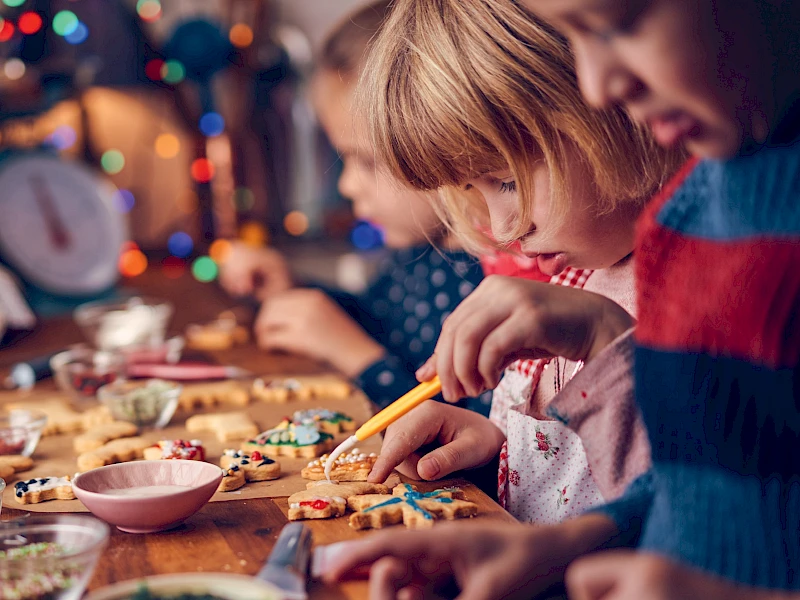 Mehrere Kinder sitzen an einem Tisch und verzieren gemeinsam Weihnachtsplätzchen mit Zuckerguss und Streuseln, im Hintergrund leuchten bunte Lichter und sorgen für eine warme, weihnachtliche Atmosphäre.
