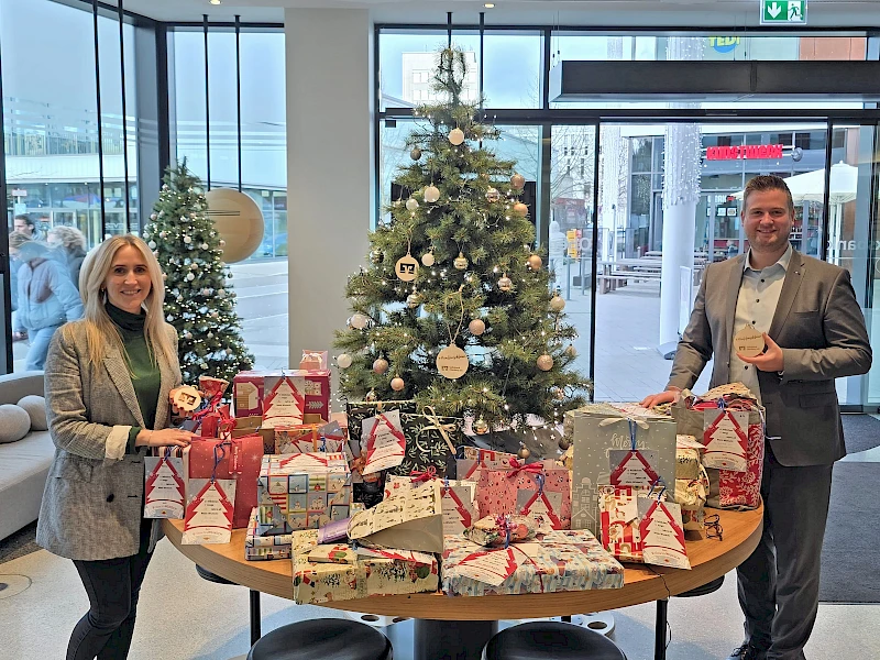 Miriam Elsner und Forum-Geschäftsstellenleiter Justin Wagner stehen im Forum Gummersbach neben einem geschmückten Weihnachtswunschbaum mit vielen verpackten Geschenken aus der Wunschbaumaktion der Volksbank Oberberg.