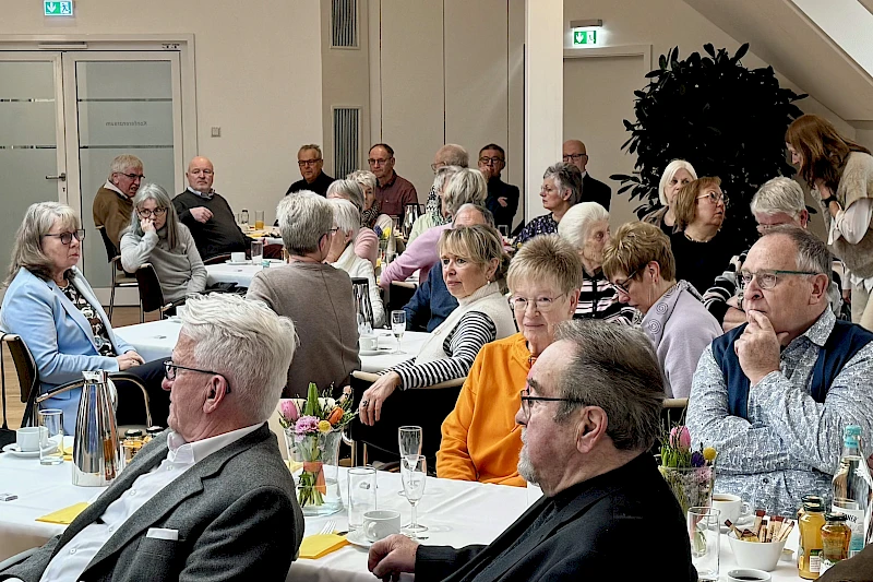 Mehrere ältere Frauen und Männer sitzen in einem hellen Konferenzraum an gedeckten Tischen mit Kaffee, Wasser und Blumenvasen und hören aufmerksam zu.