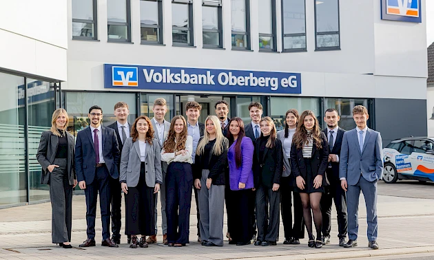 Gruppenfoto vor der Hauptgeschäftsstelle der Volksbank Oberberg in Wiehl. Links steht Ausbildungsleiterin Sarina Scherbaum, daneben die 15 neuen Bankkaufleute in Businesskleidung, im Hintergrund das Banklogo und die Fassadenfront.