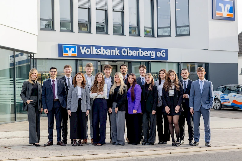 Gruppenfoto vor der Hauptgeschäftsstelle der Volksbank Oberberg in Wiehl. Links steht Ausbildungsleiterin Sarina Scherbaum, daneben die 15 neuen Bankkaufleute in Businesskleidung, im Hintergrund das Banklogo und die Fassadenfront.