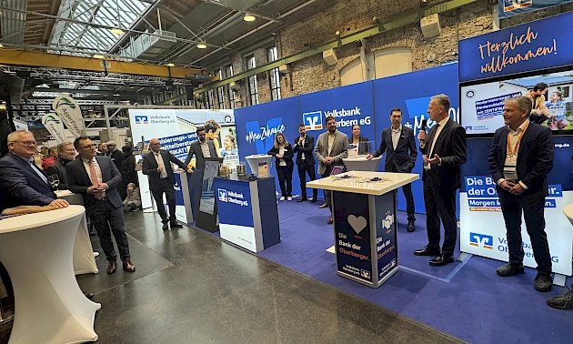 Eröffnungsszene am neuen Messestand der Volksbank Oberberg in der Halle 32. Mehrere Personen stehen vor der blau gebrandeten Standwand, ein Redner spricht ins Mikrofon, Besucherinnen und Besucher verfolgen den Auftakt.