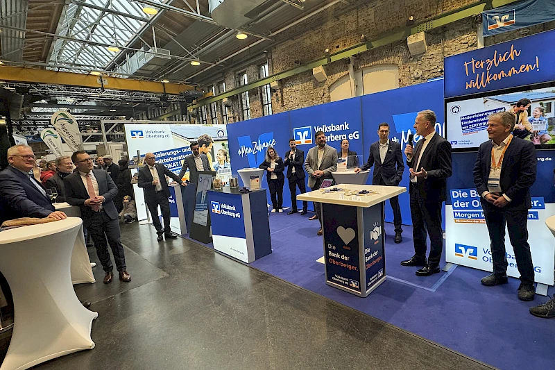 Eröffnungsszene am neuen Messestand der Volksbank Oberberg in der Halle 32. Mehrere Personen stehen vor der blau gebrandeten Standwand, ein Redner spricht ins Mikrofon, Besucherinnen und Besucher verfolgen den Auftakt.