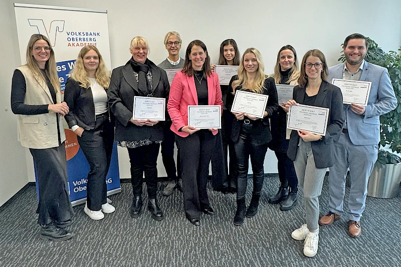 Gruppenfoto der erfolgreichen Absolventinnen und des Absolventen der Aufbauqualifizierung „Volksbank Oberberg Assistent“ mit ihren Zertifikaten, gemeinsam mit den Trainerinnen Lisa Dross und Mara Mischke in den Räumen der Volksbank Oberberg Akademie.