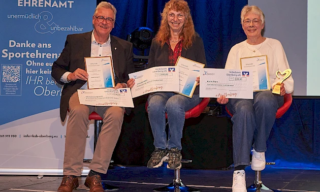 Drei ausgezeichnete Ehrenamtliche sitzen auf der Bühne und halten ihre Urkunden sowie symbolische Schecks in den Händen. Von links nach rechts: Harald Langusch, Claudia Kiel und Karin Marx, die zusätzlich eine goldene Herz-Trophäe hält. Im Hintergrund ist