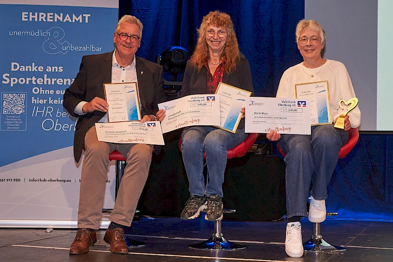 Drei ausgezeichnete Ehrenamtliche sitzen auf der Bühne und halten ihre Urkunden sowie symbolische Schecks in den Händen. Von links nach rechts: Harald Langusch, Claudia Kiel und Karin Marx, die zusätzlich eine goldene Herz-Trophäe hält. Im Hintergrund ist