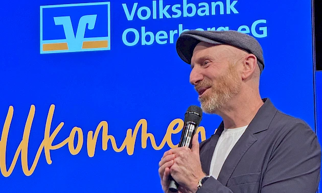 Martin Schopps steht auf der Bühne des Mitgliederabends der Volksbank Oberberg in Waldbröl und spricht mit Mikrofon vor blau beleuchtetem Bühnenhintergrund.