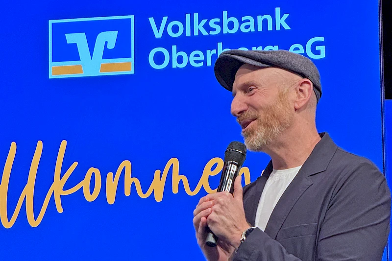 Martin Schopps steht auf der Bühne des Mitgliederabends der Volksbank Oberberg in Waldbröl und spricht mit Mikrofon vor blau beleuchtetem Bühnenhintergrund.