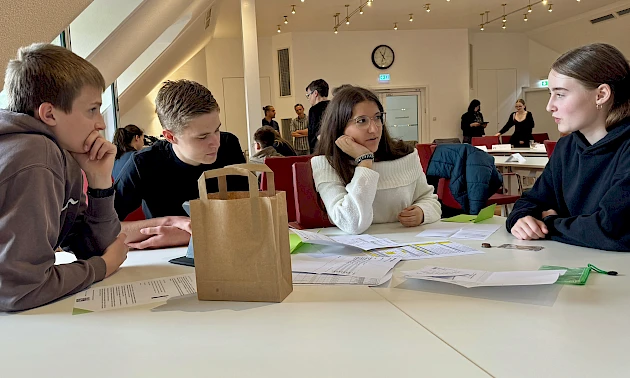 Mehrere Schülerinnen und Schüler arbeiten bei einem Workshop der Volksbank Oberberg in gemischten Kleingruppen gemeinsam an Unterlagen und Aufgaben des Mystery Games zur Buchhaltung.