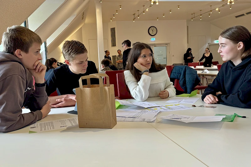 Mehrere Schülerinnen und Schüler arbeiten bei einem Workshop der Volksbank Oberberg in gemischten Kleingruppen gemeinsam an Unterlagen und Aufgaben des Mystery Games zur Buchhaltung.