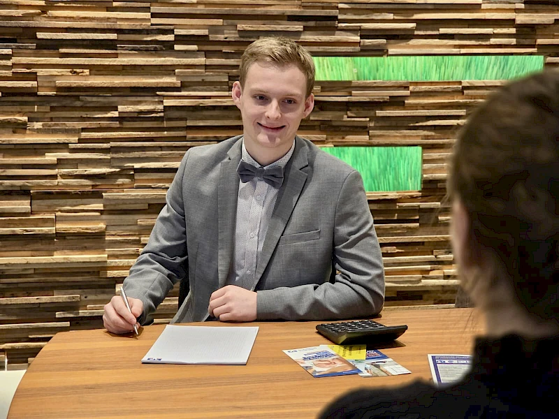 Luca Liesenberg sitzt in einer Geschäftsstelle der Volksbank Oberberg an einem Beratungstisch und führt ein Gespräch mit einer Kundin.