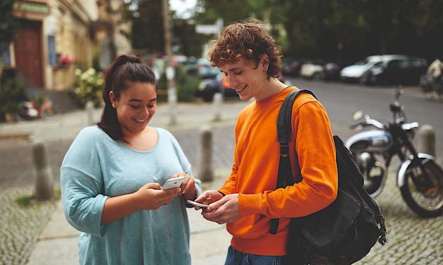 Zwei Jugendliche stehen mit Smartphones in der Hand im Freien und beschäftigen sich mit digitalen Anwendungen. Das Bild dient als Symbolfoto für Finanzbildung und den sicheren Umgang mit digitalen Zahlungsmöglichkeiten.
