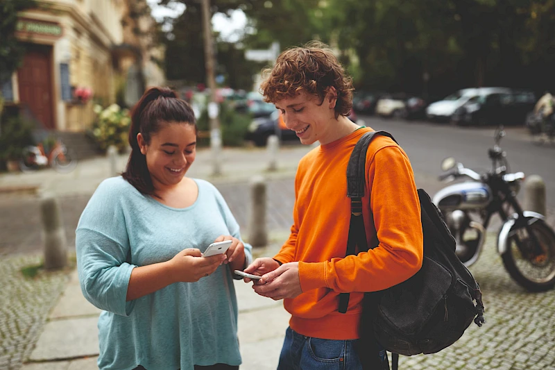 Zwei Jugendliche stehen mit Smartphones in der Hand im Freien und beschäftigen sich mit digitalen Anwendungen. Das Bild dient als Symbolfoto für Finanzbildung und den sicheren Umgang mit digitalen Zahlungsmöglichkeiten.