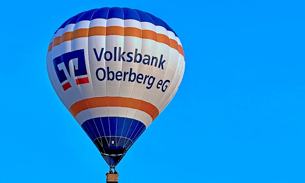 Der Heißluftballon der Volksbank Oberberg schwebt vor blauem Himmel. Auf der weißen Ballonhülle sind das Logo und der Schriftzug der Volksbank Oberberg zu sehen.