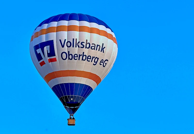 Der Heißluftballon der Volksbank Oberberg schwebt vor blauem Himmel. Auf der weißen Ballonhülle sind das Logo und der Schriftzug der Volksbank Oberberg zu sehen.