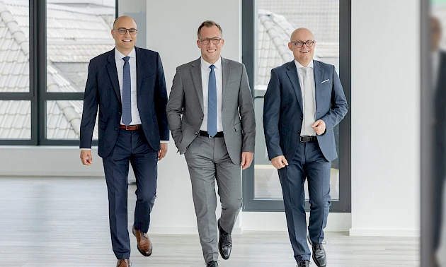Das neue Vorstandsteam der Volksbank Oberberg geht gemeinsam durch einen hellen Gebäudeflur. In der Mitte ist Vorstandsvorsitzender Andreas Neumann zu sehen, rechts der stellvertretende Vorstandsvorsitzende Jörn Richling und links Vorstandsmitglied Marc G