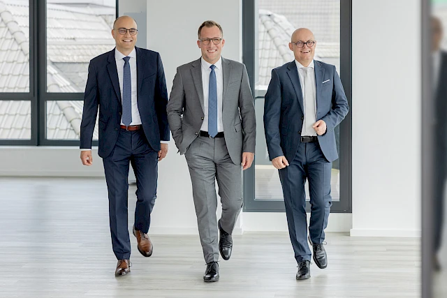 Das neue Vorstandsteam der Volksbank Oberberg geht gemeinsam durch einen hellen Gebäudeflur. In der Mitte ist Vorstandsvorsitzender Andreas Neumann zu sehen, rechts der stellvertretende Vorstandsvorsitzende Jörn Richling und links Vorstandsmitglied Marc G