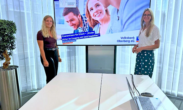 Lisa Dross und Mara Mischke stehen vor einem Bildschirm mit einer Präsentation zur Workshop-Reihe „Finanzwissen für Eltern“ in der Geschäftsstelle Waldbröl und informieren Eltern über den verantwortungsvollen Umgang mit Geld.