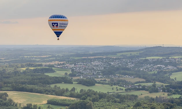 Der Heißluftballon D-OVOB der Volksbank Oberberg fliegt über die hügelige Landschaft des Oberbergischen Kreises. Im Hintergrund sind Ortschaften, Felder und Wälder unter einem Abendhimmel mit aufziehenden Regenwolken zu sehen.