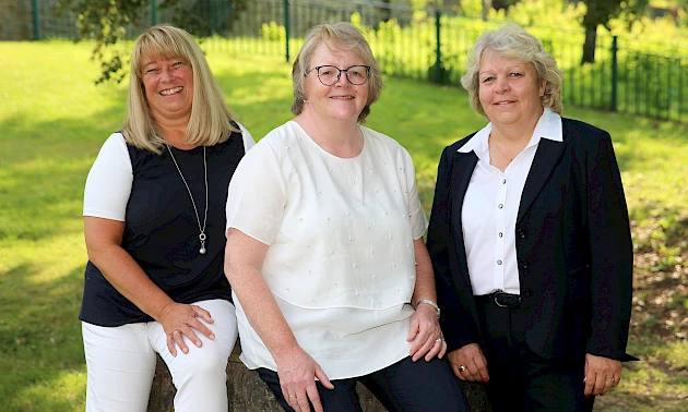 Claudia Koschwitz, Anette Nolte und Anja Ufer sitzen und stehen lächelnd auf einem Baumstamm im Wiehlpark in der Nähe der Hauptstelle Wiehl.