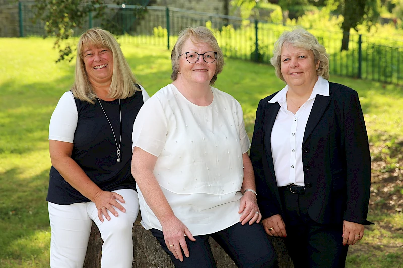 Claudia Koschwitz, Anette Nolte und Anja Ufer sitzen und stehen lächelnd auf einem Baumstamm im Wiehlpark in der Nähe der Hauptstelle Wiehl.