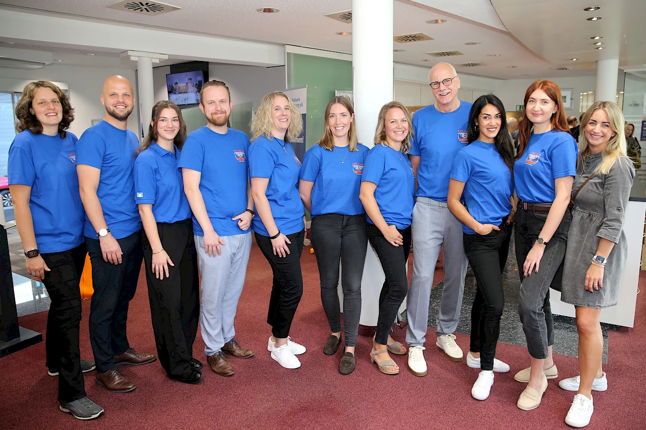 Gruppenfoto des Teams der Gesch&auml;ftsstelle Bielstein in den R&auml;umen der Volksbank Oberberg. Die Mitarbeitenden stehen nebeneinander und tragen &uuml;berwiegend blaue Jubil&auml;umsshirts zum 125-j&auml;hrigen Bestehen des Standorts. Im Hintergrund sind Teile der Gesch&auml;fts