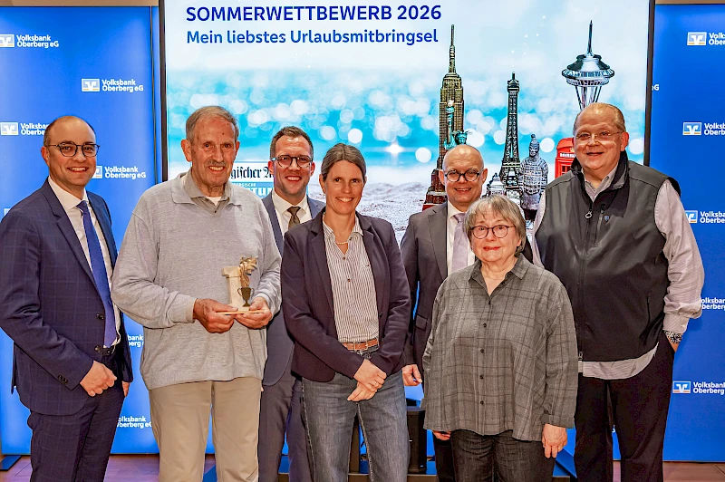 Die drei Erstplatzierten des Sommerwettbewerbs 2026 posieren bei der Preisverleihung gemeinsam mit Vertretern der Volksbank Oberberg und der Oberbergischen Volkszeitung vor der Wettbewerbswand.
